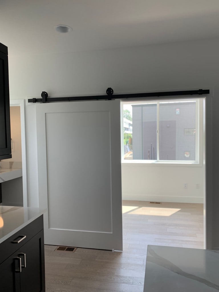 Kitchen with a sliding barn door on a black rail opening to a bright finished room by Artbrick